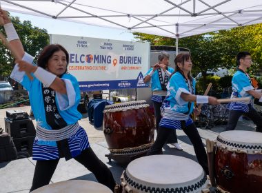 A group of drummers play large round drums on stage with a single woodwind player.